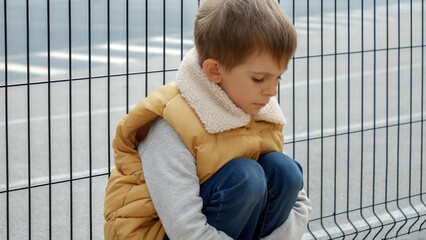 Upset lonely boy being bullied at school sitting next to metal fence at playground. Child...