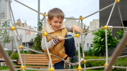 Happy cheerful boy climbing through ropes and nets on the public playground. Active child, sports and development, kids playing outdoors