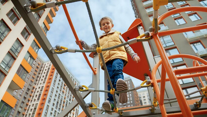 Little smiling boy crossing rope bridge between two towers on outdoor kids playground