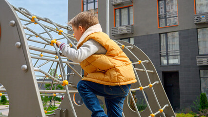 Little boy climbing up on the rope net at public children playground in the yard. Active child,...