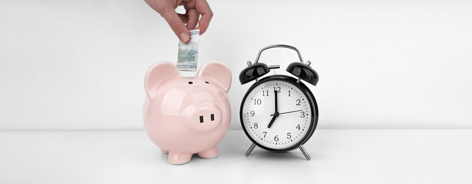 Womans hand put dollar bancnote into a piggy bank with alarm clock