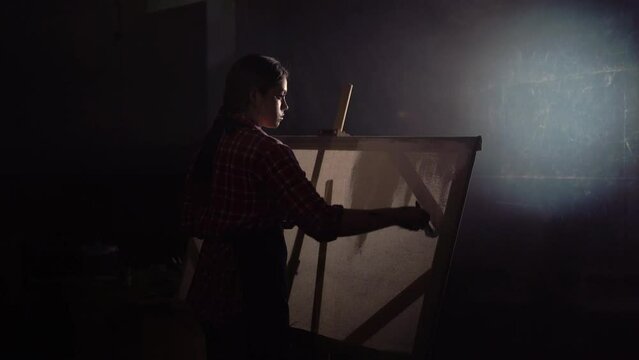 Female artist works on abstract oil painting in dark creative studio, She creates modern masterpiece with paint brush. Large canva stands on easel, light.