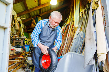 An elderly man with beard in work clothes assembles a wheelbarrow in a workshop.