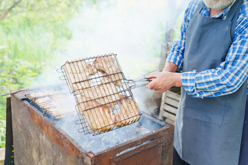 an elderly man with a beard in work clothes cooks meat steaks on the grill 