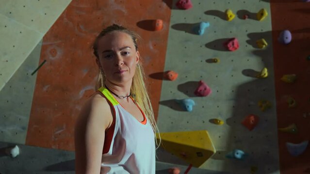 A young female climber or climber stands in front of a climbing wall indoors in the evening and turns her head to the camera. There is a place for infographics. High quality 4k footage
