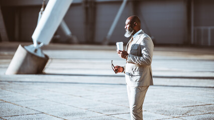 A dapper black businessman in a beige suit is captured in profile against a stunning tensile structure. He holds a cup of coffee in one hand and a pair of glasses in the other, very urban elegantly
