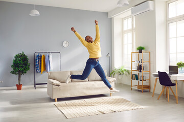 Happy young Afro American woman in yellow jumper and jeans spends weekend at home, feels full of energy and jumps up in living room with sofa, rug, clothes rack, plants, shelves and desk with laptop