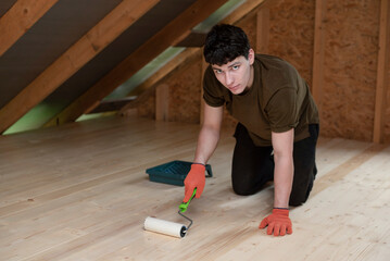 A young guy is varnishing a new wooden floor with a protective gloss varnish and roller, this is a new job for the guy and he is diligently applying the varnish to floor in summer house