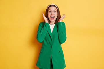 Extremely amazed woman wearing green jacket standing screaming with excitement pleasant surprise raised arms isolated over yellow background.