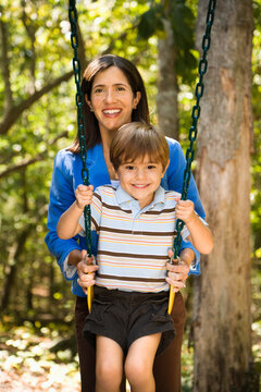 Hispanic mother pushing son on swing and smiling at viewer.