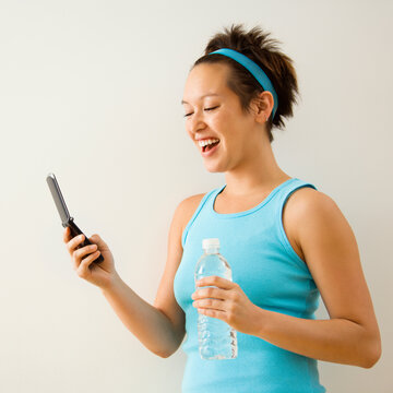 Young woman in fitness clothing holding bottled water and smiling at cellphone.