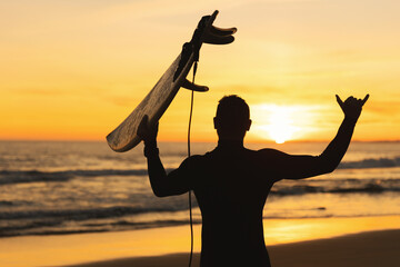Silhouette of an athletic man holding a surfboard showing shaka at sunset