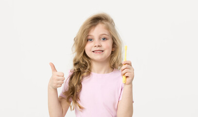 A cute blond child girl on a white background is holding a toothbrush near her face. She smiles broadly and shows the class sign with her finger. Children's dentistry and oral hygiene concept
