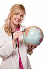 Fototapeta premium Attractive friendly smiling blond lady doctor in white lab coat and pink scrubs holding a stethoscope listening on a globe on white