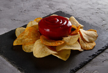 Side view of heap of potato chips with red dipping sauce on black slate serving board on grey...