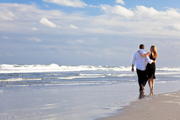 A young man and woman arms around each other walking as a romantic couple on a beach