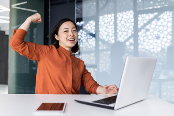 A young Asian woman student studying using a laptop in a co-working space. Satisfied with the test result, the examinee shows a hand gesture of strength and success. Smiling, she looks at the camera.