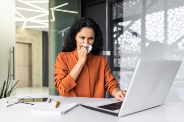 A young muslim woman works in the office on a laptop, sits at a table and feels bad. He has a runny nose, allergies, a virus. He wipes his nose with a napkin, sneezes and coughs, needs treatment.