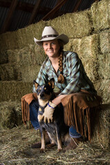 Attractive young woman wearing a white cowboy hat. She is sitting on hay and smiling while holding an Australian Shepherd. Vertical shot.