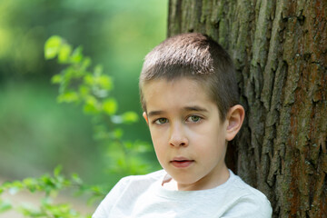 small blue-eyed boy in a gray t-shirt leans his back against a tree and looks directly into the camera, with a green blurred background