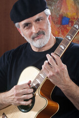 Portrait of a middle-aged man wearing a black beret and t-shirt and playing an acoustic guitar. He is looking at the camera. Vertical shot.