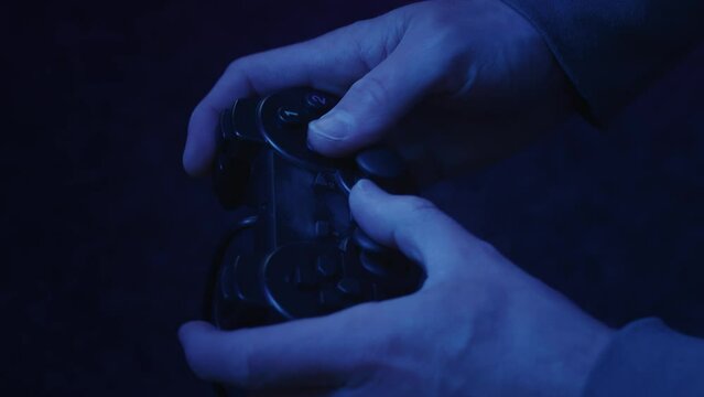 A video gamer rapidly shifts thumbsticks and hammers at buttons on an unbranded generic game controller illuminated only by a pink and blue flickering TV screen.