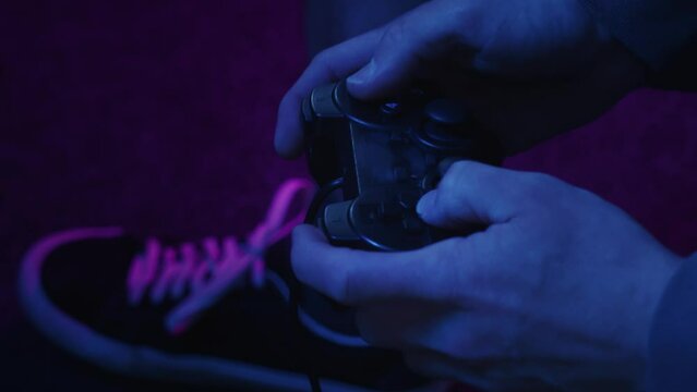 A video gamer rapidly shifts thumbsticks and hammers at buttons on an unbranded generic game controller illuminated only by a pink and blue flickering TV screen.