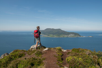 Fototapeta premium Caucasian tourist with a red backpack standing on top of Sukkertoppen mountain in Ålesund during summer, enjoying the beautiful view of the surrounding islands and ocean. Norway, day, enjoyment