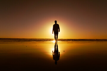 Silhouette of a young man walking on the beach backlit by setting sun and his reflection in the wet sand