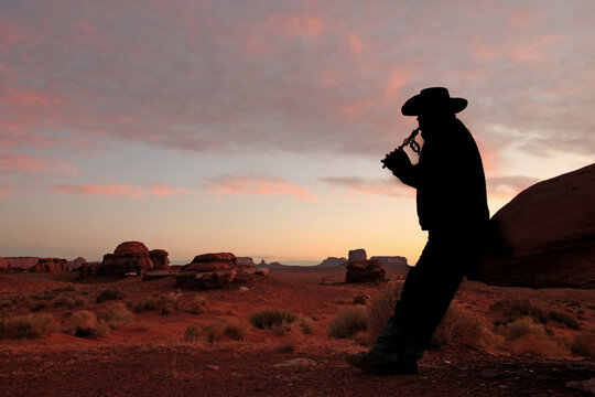 Mid adult male wearing cowboy hat and playing the flute. He is full length viewable, looking away from the camera and in silhouette. Horizontally framed shot.