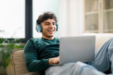 Arabic Guy Typing And Websurfing On Laptop Wearing Earphones Indoors