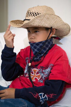 Young asian boy sitting on a white sofa wearing a cowboy hat and a bandana with finger on the brim of the hat