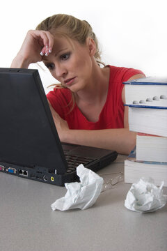 Young woman sitting in front of laptop beside a pile of thick textbooks and crumpled paper with hand on forhead looking stressed