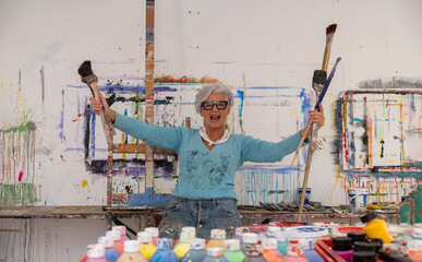 older gray haired mature happy laughing artist woman with glasses and big paintbrushes in her hands, in studio