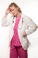 Attractive blond lady doctor in white lab coat and pink scrubs with a stethoscope holding forehead with a pained expression