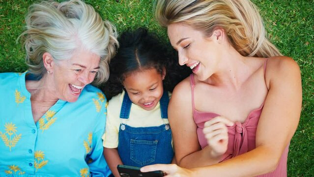 Overhead shot of smiling multi-generational female family lying on grass in garden at home together taking selfie on mobile phone- shot in slow motion