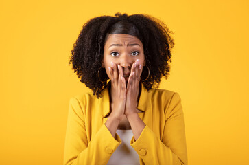 Shocked afraid black woman covering her mouth with hands, looking at camera, isolated on yellow...