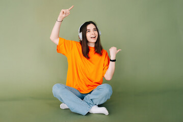 Portrait of a joyful woman wearing headphones, expressing enthusiasm and approval while pointing both thumbs towards empty space, isolated on a vibrant green background.