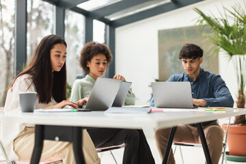 Three Multiethnic Coworkers Working Using Laptops At Table In Office