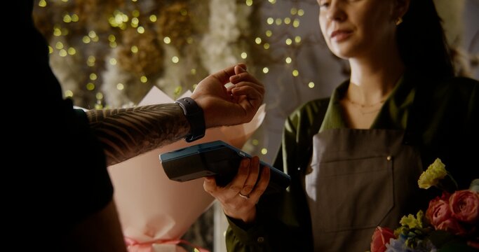 Man buys spring bouquet in flower shop and pays for purchase with contactless payment using smart watch. Female florist sells fresh flowers to male customer. Floristry and entrepreneurship concept.