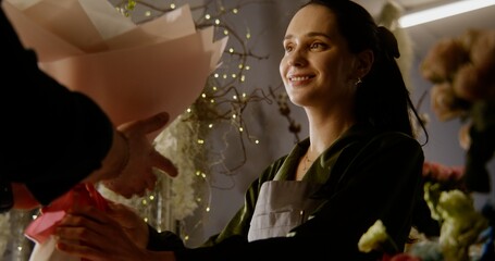 Female florist, seller gives payment terminal to male buyer. Man pays for flowers bouquet in wrapping paper with contactless payment using phone in flower store. Floristry, small business. Close up.
