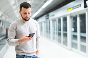 Interested young man browsing messages on phone while waiting for subway train, standing at station..