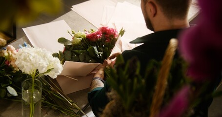 Male professional florist, entrepreneur, seller wraps fresh flowers bouquet in wrapping paper for customer in flower shop. Concept of floristry, floral small business and entrepreneurship. High angle.