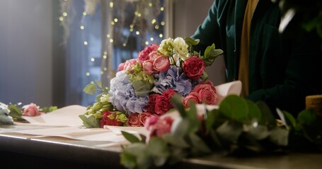 Close up shot of professional male florist wrapping beautiful bouquet of fresh flowers using wrapping paper. Process of working in flower shop. Concept of retail floral business and entrepreneurship.