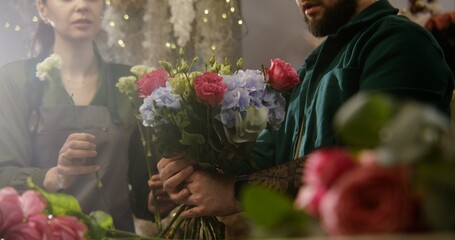 Fototapeta premium Close up shot of professional florist entrepreneur adding fresh flowers to bunch in floral shop. Woman helps colleague to collect beautiful bouquet. Concept of floral business and entrepreneurship.