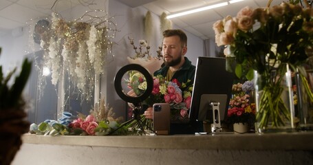Male florist collects bouquet of flowers in flower shop, records video for blog using ring lamp, smartphone and digital tablet. Concept of floristry, retail floral small business and entrepreneurship.