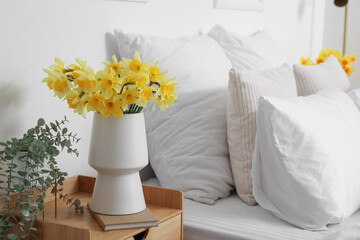 Vase with blooming narcissus flowers and houseplant on bedside table