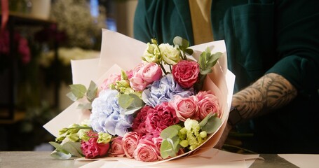 Male florist wraps beautiful bouquet, uses wrapping paper. Colleague with vase of flowers walks at background. Process of working in flower shop. Floral business and entrepreneurship concept. Close up