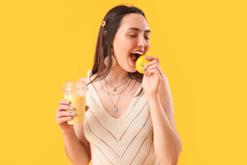 Young woman with glass of smoothie eating lemon on yellow background