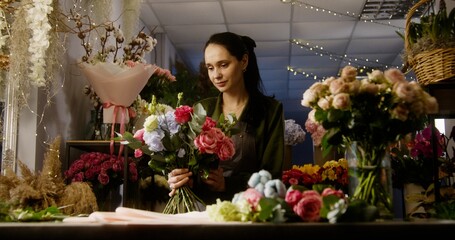 Professional female florist keeps in hands beautiful bouquet, smiles and looks at camera. Vases with plants and fresh flowers stand at background. Retail floral business and entrepreneurship concept.